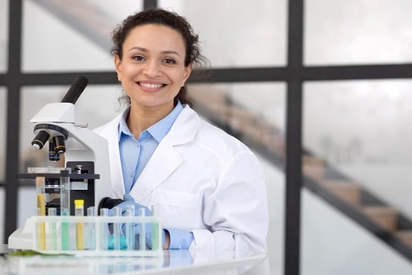 Млада жена учен с тъмна къдрава кода, стои усмихната до микроскоп