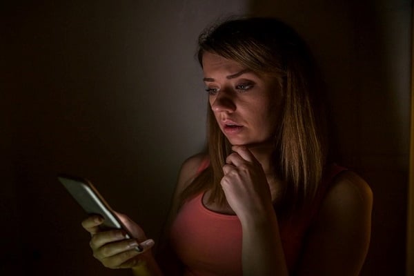 Млада жена с оранжев потник, която гледа екрана на телефон в тъмна стая