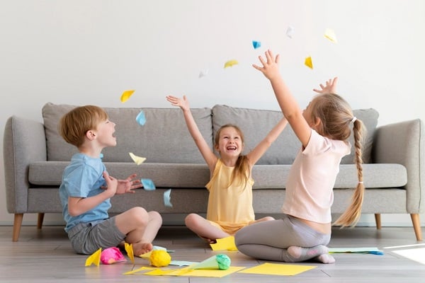 Три деца, седнали на пода, които подхвърлят шарени хартиени самолетчета във въздуха