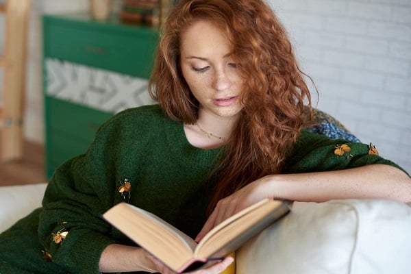 Червенокоса млада жена с лунички и зелен пуловер, която чете книга