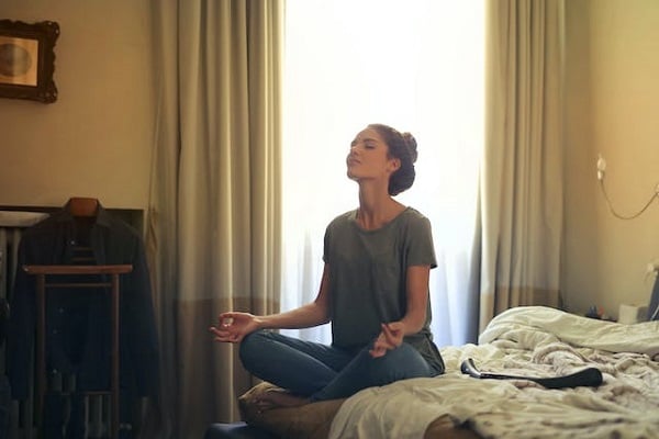 Жена със зеленикава тениска, която е седнала на легло и медитира