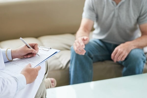 Мъж със сива тениска и дънки, който е седнал на диван и разговаря с лекар