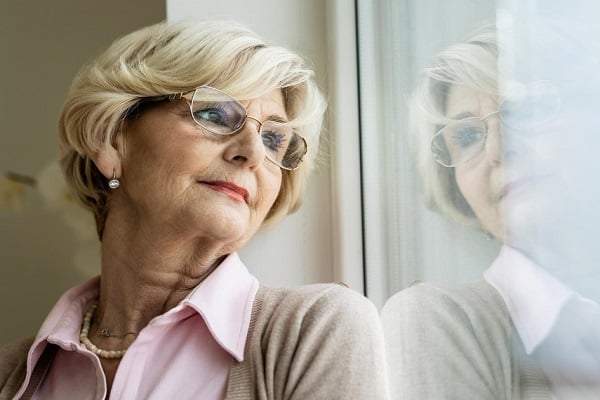 Възрастна жена с руса къса коса и очила, която гледа през прозорец